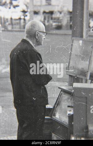 Fine anni '70 vintage nero e bianco stile di vita fotografia di un uomo anziano fuori per la sua passeggiata mattutina acquistare un giornale dalla locale edicola. Foto Stock