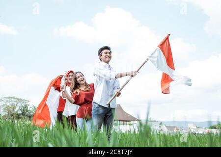 L'uomo asiatico tiene e solleva bandiera indonesiana mentre segue dietro il suo amico indossando attributi rossi e bianchi nel campo di riso con uno sfondo di paesaggi naturali Foto Stock