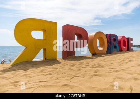 Iscrizione multicolore Yarovoe in lingua russa sulla sabbia gialla sullo sfondo del lago e dei turisti. Yarovoe nome della città di villeggiatura nel Foto Stock