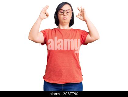 Donna bruna con sindrome di Down indossare abiti e occhiali casual rilassarsi e sorridere con gli occhi chiusi facendo meditazione gesto con le dita. yoga co Foto Stock