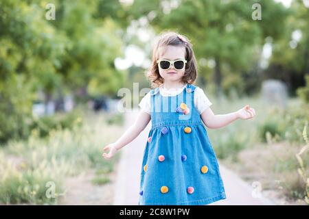 Elegante bambina di 2-3 anni con abiti in denim e occhiali da sole all'aperto nel parco. Guardando la fotocamera. Stagione estiva. Foto Stock