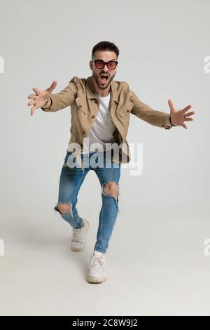 giovane ragazzo casual sconvolto in giacca indossare occhiali da sole, urlare e correre, prendere e posare su sfondo grigio Foto Stock