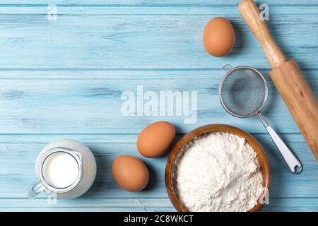 Ingredienti per la cottura, il latte, le uova, la farina di grano e utensili da cucina su sfondo di legno blu, vista dall'alto Foto Stock