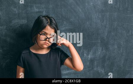 Ritorno a scuola. Ragazza piccola seria in occhiali pensando di toccare la testa con un dito vicino a lavagna. Bambino della scuola elementare. Foto Stock