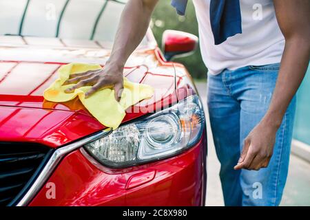 Closeup immagine tagliata di mani di giovane africano, lavando e pulendo la sua auto rossa con panno in microfibra gialla, cappuccio e fari. Auto Foto Stock