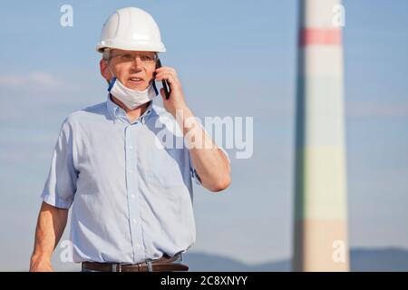 Ingegnere senior o maturo, architetto o ispettore con maschera facciale che parla sul telefono cellulare in un cantiere o in una fabbrica - fuoco selettivo Foto Stock