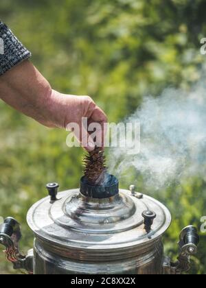 Donna mano tenere cono abete vicino annata fumo Samovar, la caldaia tradizionale russa acqua. Fuoco samovar all'aperto. Verticale. Spazio di copia per testo o disegno. Foto Stock