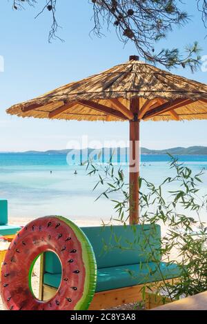 Salotto in legno con ombrellone, panca e anello gonfiabile all'ombra della pineta sulla spiaggia paradisiaca che si affaccia sul mare e il cielo blu. Foto Stock