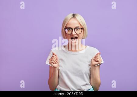 Scioccata donna sorpresa accoppia i pugni con felicità, apre la bocca ampiamente in stupore, posa isolato contro sfondo viola con copyspace Foto Stock