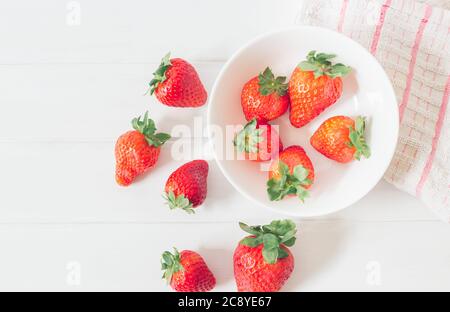 Vista dall'alto di fragole fresche su un tavolo di legno bianco rustico, all'interno di una ciotola pronta a mangiare come cibo biologico e sano, accanto a un panno con striscia rossa Foto Stock