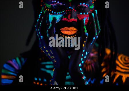 Ritratto concettuale di giovane africano dipinto in colori UV fluorescenti incandescenti, l'uomo tocca il viso, con gli occhi chiusi. Make-up, concetto di body art, is Foto Stock