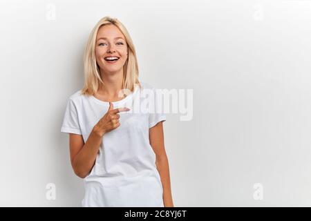 Felice giovane donna di charme ha un'espressione gioiosa, punta con le dita da parte, indossa una t-shirt casual, isolato su sfondo bianco con spazio vuoto. cop Foto Stock