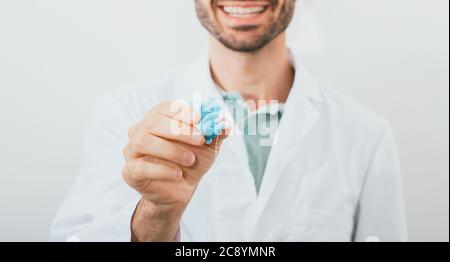 Un audiologo positivo che tiene in mano ha modellato gli auricolari individuali per il suo paziente, primo piano. Soluzione per l'udito Foto Stock