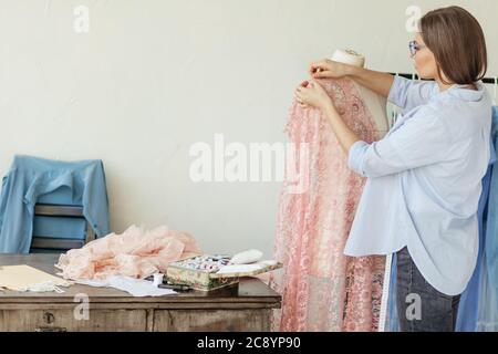 Giovane marinaio caucasico inserisce gli aghi in un abito da sposa in pizzo vestito di manichino. Creazione di abiti di moda in studio di moda. Foto Stock