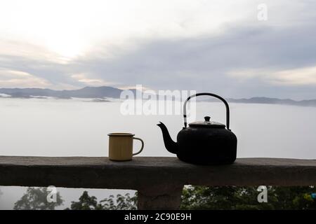 Chiudete una tazza di caffè caldo in lattina gialla con il vecchio bollitore e la nebbia sullo sfondo della montagna al mattino. Foto Stock