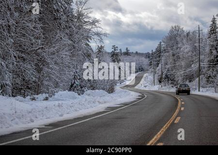 Un'automobile più vecchia sta guidando giù una strada tortuosa di paese nell'inverno, con gli alberi di foresta su entrambi i lati della strada rivestiti in uno strato spesso di neve fresca. Foto Stock