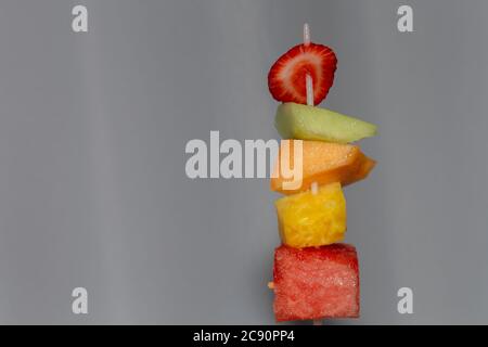 Uno spiedino di plastica ha tagliato pezzi di frutta succosa: Fragola, melone di miele, cantalupe, ananas e anguria, ed è tenuto in su prima di un backgr bianco Foto Stock