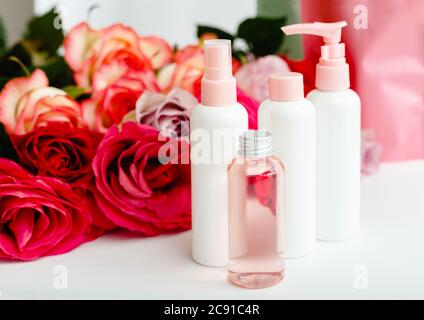 Bottiglie cosmetiche in plastica, siero, sapone, olio su sfondo floreale da tavola bianco. Fiore rosso rosa rose naturale prodotto di bellezza organico. Centro benessere, cura della pelle Foto Stock