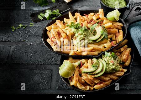 Due padelle con porzioni di patatine fritte croccanti e condite e guacamole con pera di avocado a fette su un bancone piastrellato nero con copyspace Foto Stock