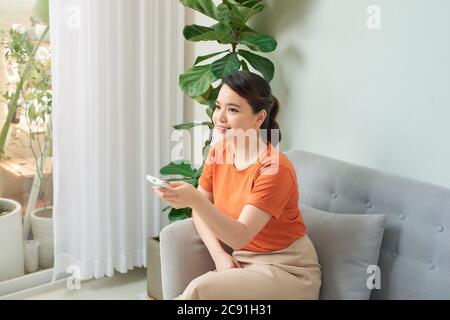 Felice giovane donna che accende il condizionatore d'aria con telecomando a casa Foto Stock