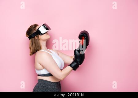 vista laterale di una ragazza in sovrappeso con visore vr e guanti da boxe che guardano in rosa Foto Stock