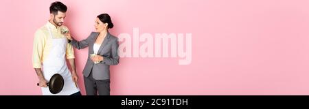 intestazione di web site di businesswoman che mette i soldi in tasca dell'uomo bello in grembiule che tiene la padella di frittura sul rosa Foto Stock