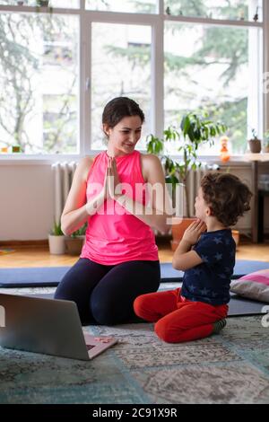 Giovane madre e bambino che fa la posa di Namaste mentre esercita lo yoga A casa davanti al notebook Foto Stock