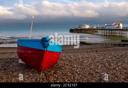 Worthing Seafront West Sussex England UK - Red & Blue pesca barca sulla spiaggia con Worthing Pier dietro al tramonto Foto Stock