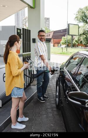 Fuoco selettivo di uomo sorridente che alimenta l'automobile vicino a moglie con il caffè per andare sulla stazione di benzina Foto Stock