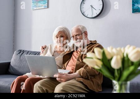 fuoco selettivo di coppia anziana felice seduta sul divano e usando il laptop vicino bouquet di tulipani Foto Stock