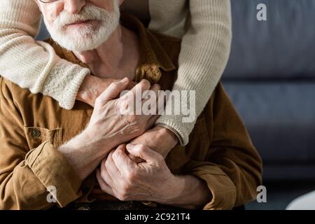 vista tagliata di donna che abbraccia il marito anziano e che tiene le mani Foto Stock