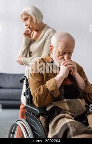 fuoco selettivo di moglie anziana triste in piedi dietro disabile, uomo malato seduto in sedia a rotelle con le mani piegate Foto Stock