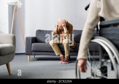 fuoco selettivo della donna disabile in sedia a rotelle e dell'uomo anziano, disprezzato seduto sul divano e coprente la testa con le mani Foto Stock