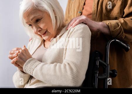 vista ritagliata del marito che tocca le spalle della donna anziana che piange mentre si siede in sedia a rotelle Foto Stock