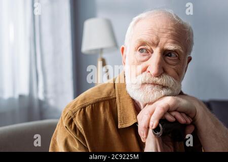 uomo anziano e solitario che guarda via mentre si siede e si appoggia su bastone da passeggio Foto Stock