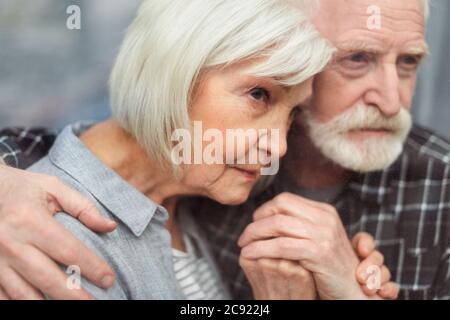 uomo anziano premuroso che tiene le mani di moglie, ammalato nella demenza, mentre guardando attraverso la finestra Foto Stock