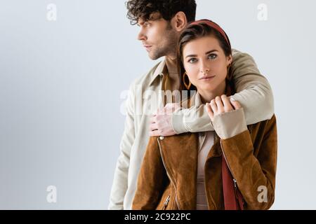 ragazza attraente e alla moda che guarda la macchina fotografica mentre l'uomo bello abbracciare la sua isolata su grigio Foto Stock