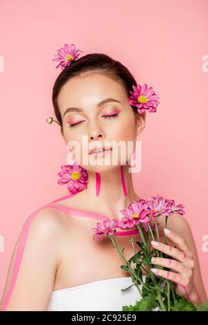 bella donna con occhi chiusi, linee rosa sul corpo e fiori in capelli che tengono bouquet isolato su rosa Foto Stock