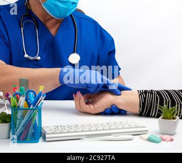 medico in una uniforme blu e una maschera usa e getta misura il polso della donna sulla sua mano destra, il concetto di ricevere e consultare i pazienti, medico Foto Stock