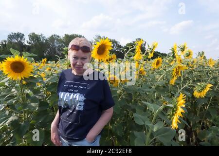 Donna matura in un campo con girasoli, sorridente, occhiali da sole sono sollevati sulla fronte Foto Stock