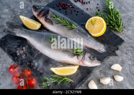 Pesce fresco crudo con pomodoro al limone e spezie su piatto di pietra nera con ghiaccio freddo nebbia di vapore. Pesce fresco al concetto di mercato. Foto Stock