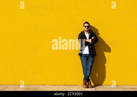 Giovane uomo bearded in piedi contro un muro giallo con le braccia incrociate Foto Stock