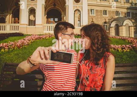 Giovane coppia amorosa che prende selfie seduto sulla panchina nel centro della città, ingannandosi e guardandosi l'un l'altro Foto Stock