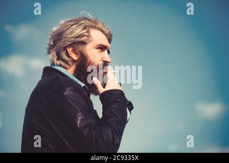 Faccia di un uomo di mezza età che fuma una sigaretta e che guarda pensieroso. Contro un cielo blu. Foto Stock