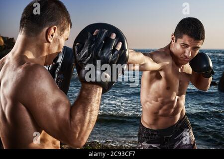 Il combattente sta addestrando le sue abilità di pugilato con un allenatore. Allenamento all'aperto in spiaggia. Pugilato, mma, wrestling, karate. Atleta giovane, forte, muscolare Foto Stock