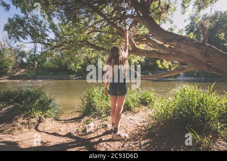 adorabile ragazza con capelli lunghi sulla riva del fiume rilassante in verde natura panoramica in sole ora d'oro ora giorno Foto Stock