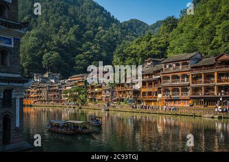 Feng Huang, Cina - Agosto 2019 : Visita di vecchie barche turistiche in legno che navigano tra le antiche case in legno palafitte costruite sulle rive del fiume del T. Foto Stock