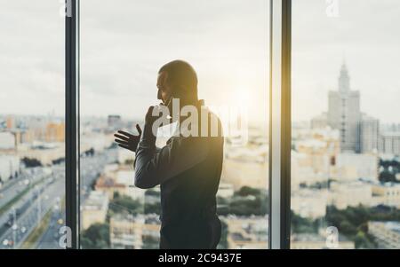 Una silhouette di un imprenditore maturo riuscito che si trova davanti ad una finestra panoramica di un alto ufficio di affari e che ha una conversazione vivace Foto Stock