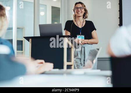 Donna d'affari sorridente in piedi sul podio e dando un discorso a persone sedute in pubblico. Imprenditrice femminile che condivide le sue esperienze in un conferenc Foto Stock