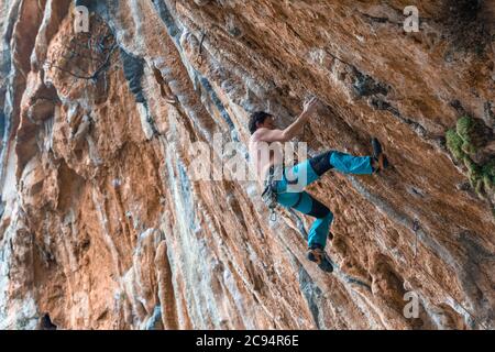 Un uomo arrampicatore di roccia in pantaloni blu brillante che arrampicano l'impegnativo percorso verticale su una parete rocciosa. Località di arrampicata Leonidio, Grecia Foto Stock